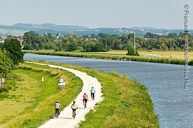 Main-Donau-Kanal bei Hirschaid (Fränkische Schweiz) Ein Fluss, an dessen grünen Ufern ein Radweg mit Radelnden vorbeiführt