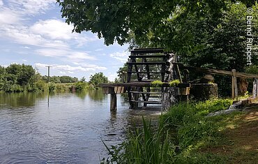 An einem Flussufer steht ein Wasserrad, daneben und dahinter sind grüne Bäume
