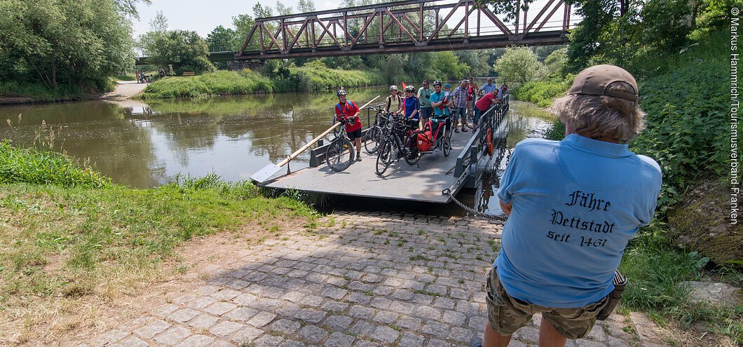 Auf einer kleinen Fähre kommt eine Gruppe Radfahrer an, ein als Fährmann erkennbarer Mann erwartet sie bereits
