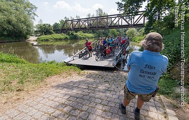 Auf einer kleinen Fähre kommt eine Gruppe Radfahrer an, ein als Fährmann erkennbarer Mann erwartet sie bereits