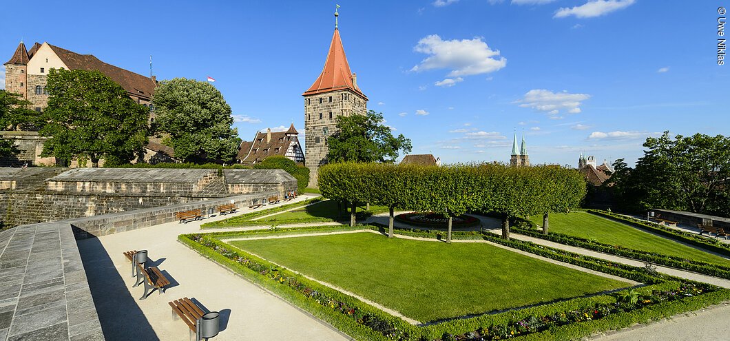 Ein gepflegter Garten mit Bänken, im Hintergrund die Türme einer Wehranlage