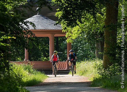 Zwei Radelnde umgeben von Grün, im Hintergrund ein Pavillon
