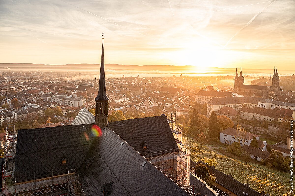 Blick über eine Kirche hinweg zur untergehenden Sonne, davor eine Stadt mit Kirchtürmen