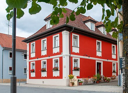Ein rotes Steinhäuschen mit weißen Fenstern, davor ein Marktplatz