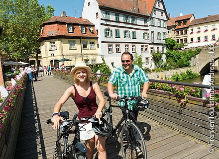 Zwei Personen schieben Ihre Räder über einen Steg, im Hintergrund sind historische Gebäude zu sehen.