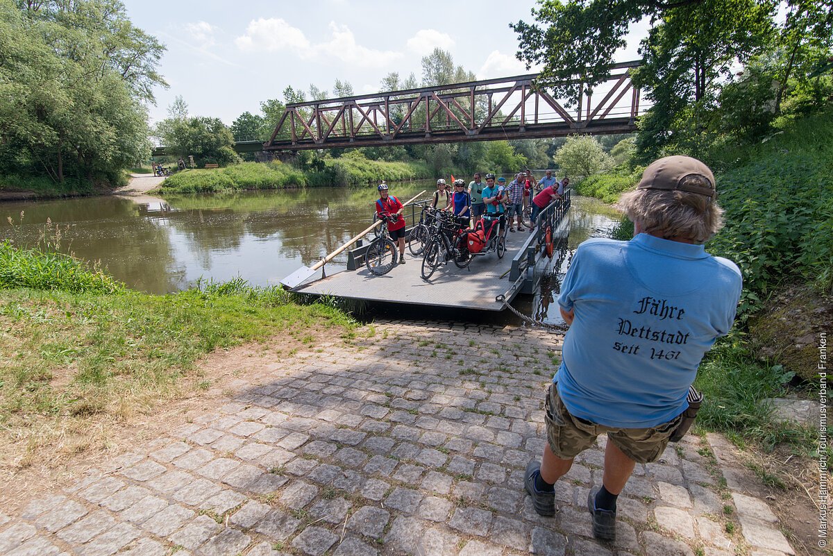 Auf einer kleinen Fähre kommt eine Gruppe Radfahrer an, ein als Fährmann erkennbarer Mann erwartet sie bereits