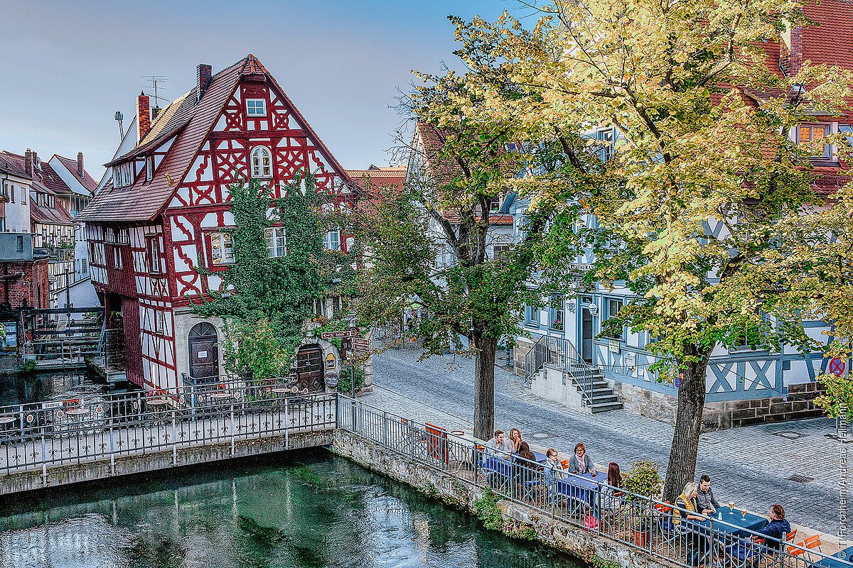 Blick auf ein Fachwerkhaus am Ufer eines Flusses, davor eine kleine Brücke