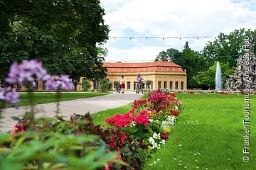 Park mit Blumenbeeten, zwei Personen mit Fahrrädern auf Weg, Gebäude und Springbrunnen im Hintergrund