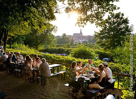 Menschen sitzen an Biergartentischen im Freien, stoßen mit Bierkrügen an, im Hintergrund Bäume und ein Dorf bei Sonnenuntergang.