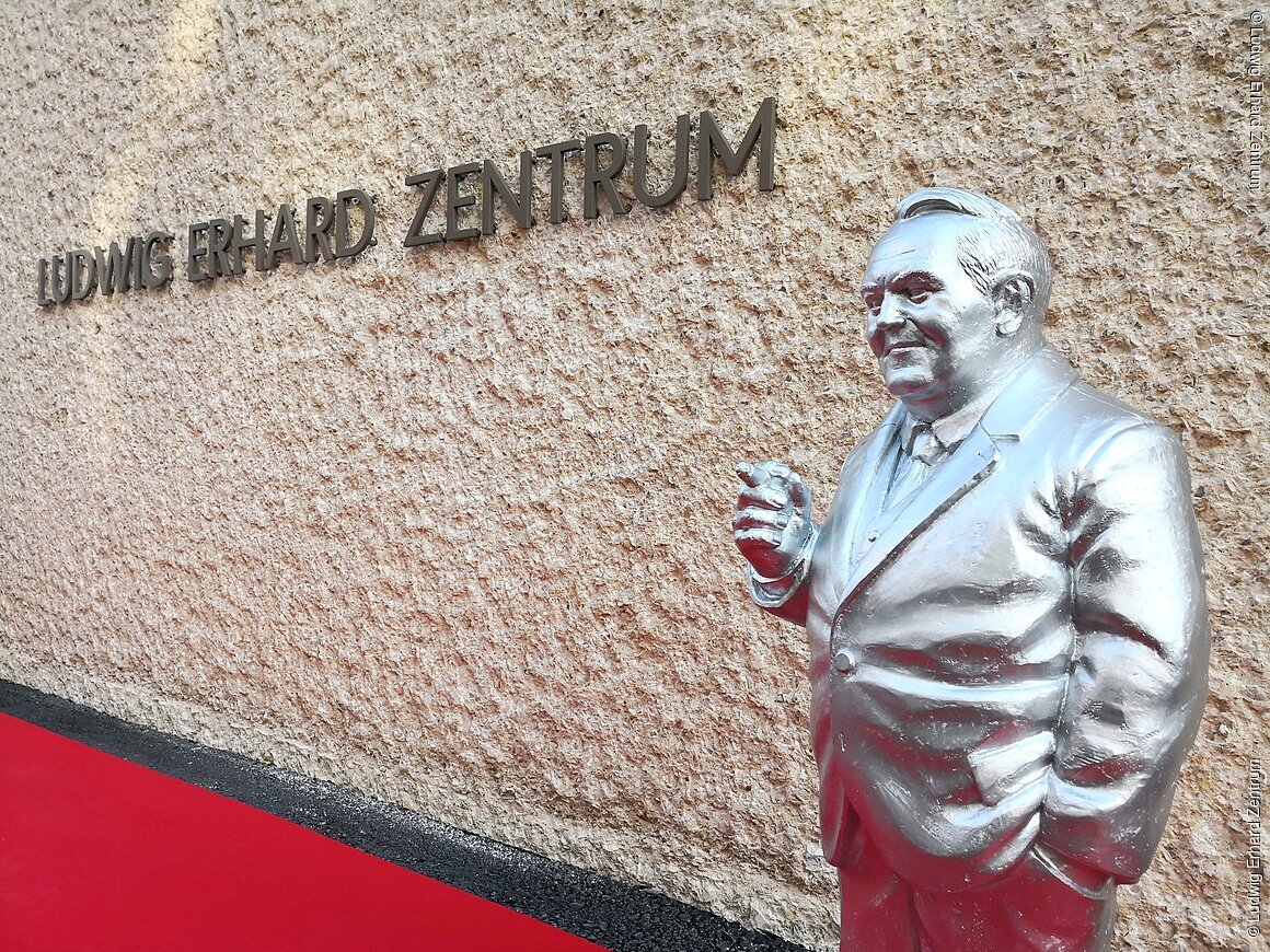 Silberne Statue eines Mannes vor einer Wand mit der Aufschrift „Ludwig Erhard Zentrum“ und rotem Teppichboden.