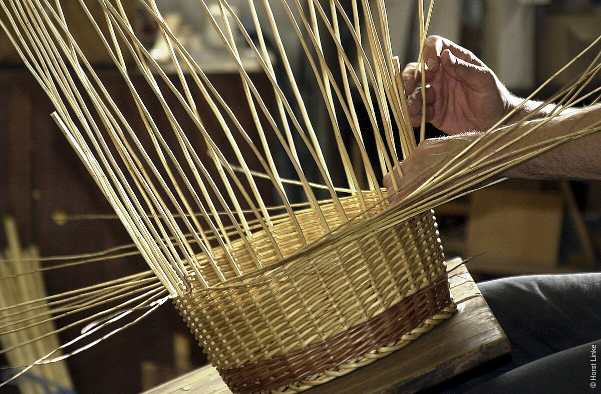 Hände flechten einen Korb aus dünnen, langen Holzstreifen auf einem Holztisch.