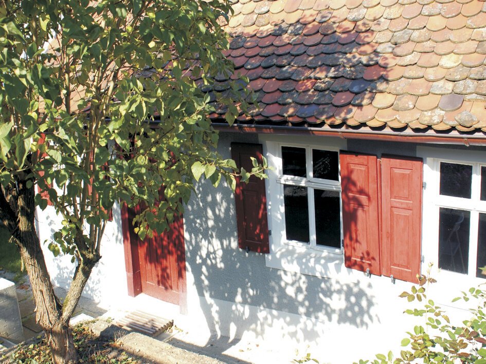 Hauswand mit zwei Fenstern und roten Fensterläden, rote Tür, Baum mit grünen Blättern davor, Dach mit Dachziegeln