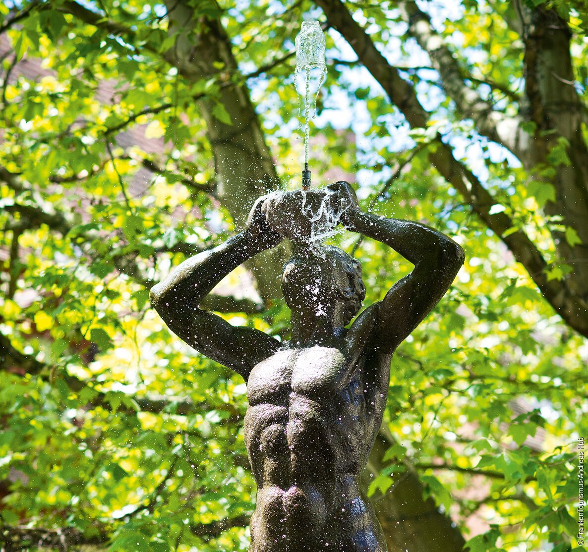 Brunnenfigur eines muskulösen Mannes mit Wasserstrahl aus dem Mund, umgeben von grünen Baumblättern.