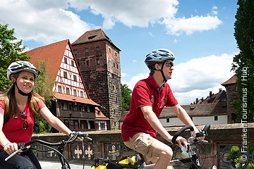 Zwei Personen mit Fahrradhelmen fahren bei Sonnenschein auf einer Straße vor Fachwerkhäusern und einem Turm.
