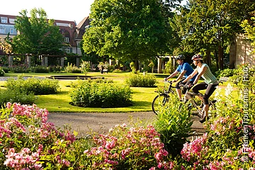 Zwei Radfahrer mit Helmen fahren auf einem Weg durch einen Park mit Blumen und Büschen bei Sonnenschein.