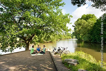 Drei Personen sitzen auf einer Decke unter einem Baum an einem Flussufer mit Fahrrädern neben sich.