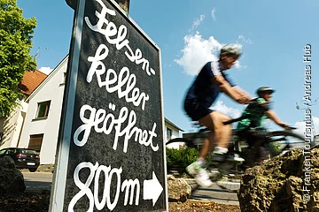 Schild mit der Aufschrift „Felsenkeller geöffnet 800 m“ und zwei verschwommene Radfahrer auf einer Straße bei Tageslicht.