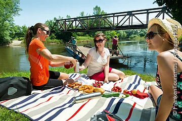 Drei Frauen sitzen auf einer Decke am Flussufer mit Picknick und Fähre im Hintergrund an sonnigem Tag.