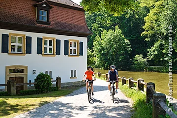 Zwei Radfahrer fahren auf einem Weg neben einem Fluss und einem weißen Haus mit blauen Fensterläden.
