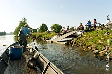 Mann in Gummihose rudert Boot auf Fluss, fünf Radfahrer sitzen und stehen am Ufer mit Fahrrädern.