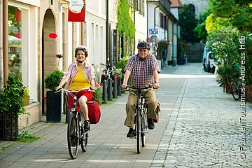Älteres Paar fährt mit Fahrrad auf einem gepflasterten Gehweg in einer Stadtstraße.