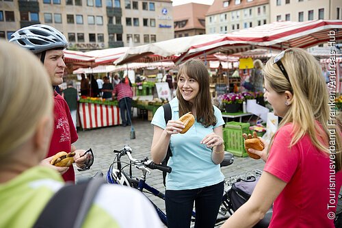 Vier Personen mit Fahrrädern essen Brötchen auf einem Markt mit Ständen und roten Markisen.