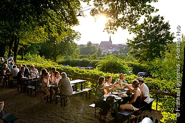 Menschen sitzen an Holztischen im Freien, trinken Bier, umgeben von Bäumen und Blick auf Dorf mit Kirche bei Sonnenuntergang.