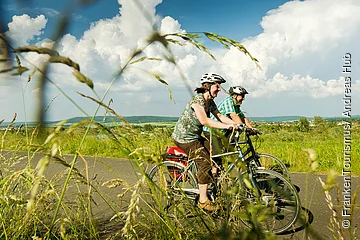 Zwei Personen mit Fahrradhelmen fahren auf einem Radweg durch eine grüne Landschaft bei bewölktem Himmel.