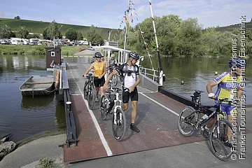 Fahrradfahrer verlassen eine Fähre auf einem Fluss, im Hintergrund Weinberge und Wohnmobile.