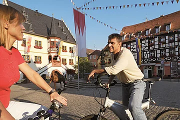 Zwei Personen auf Fahrrädern im Gespräch auf einem gepflasterten Platz mit Fachwerkhäusern und Fahnen.