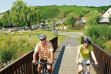 Zwei Personen fahren mit Fahrrad und Helm über eine Holzbrücke in ländlicher Umgebung bei Sonnenschein.