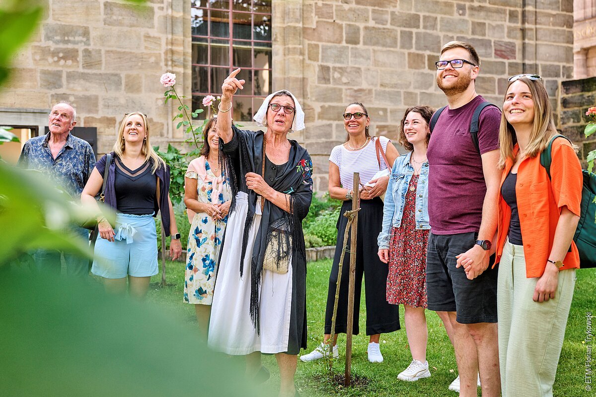 Gruppe von Menschen bei einer Führung im Freien vor einer historischen Steinmauer, eine Frau zeigt nach oben.